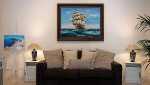 A living room with a dark brown sofa, two side tables with lamps, a framed painting of a ship on the wall, and a small easel with a painting. - Holyday Home in Historic Center very close ti Porta Vecchia Beach  (Monopoli)