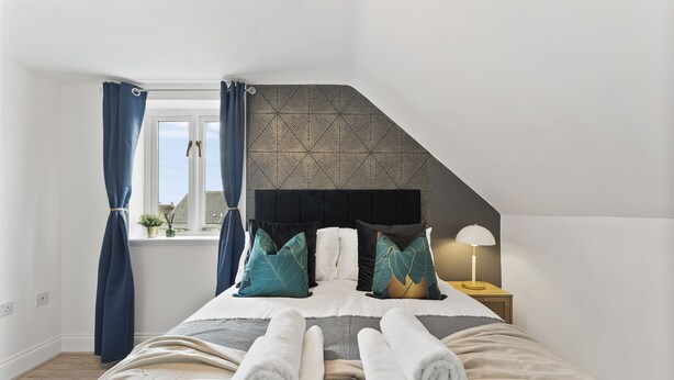 Une chambre à coucher avec un lit, des oreillers, une table de chevet, une lampe et une fenêtre avec des rideaux.