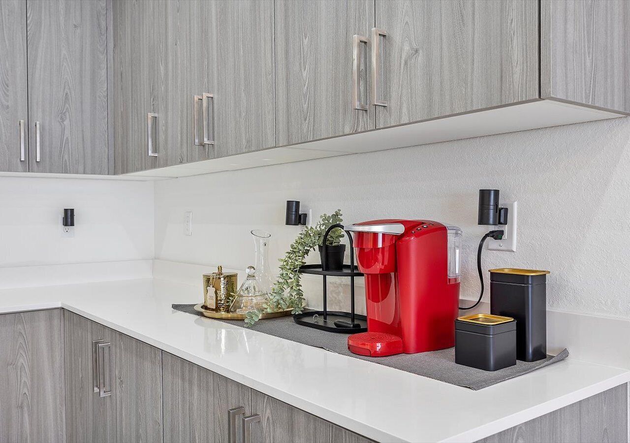 A modern kitchen with grey cabinets, a white countertop, and a red coffee maker.