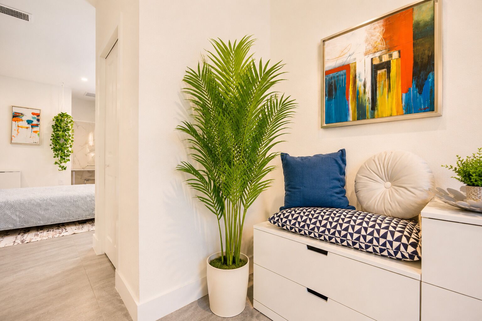 Une pièce moderne avec une grande plante en pot, un canapé agrémenté de coussins à motifs, un tableau accroché au mur et un lit à l’arrière-plan.