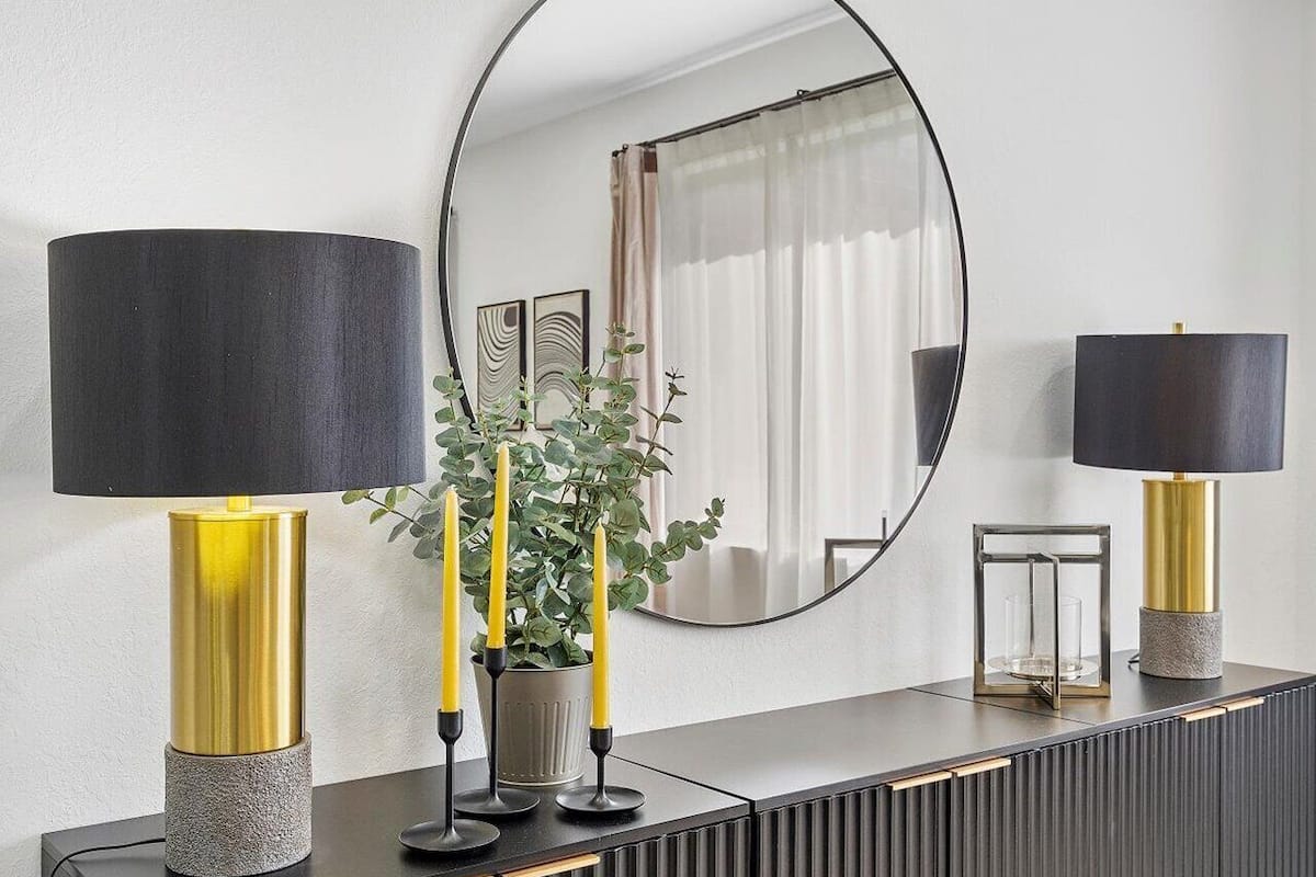 A modern bathroom with a black vanity, two gold-toned lamps, a mirror, and a potted plant with yellow candles.