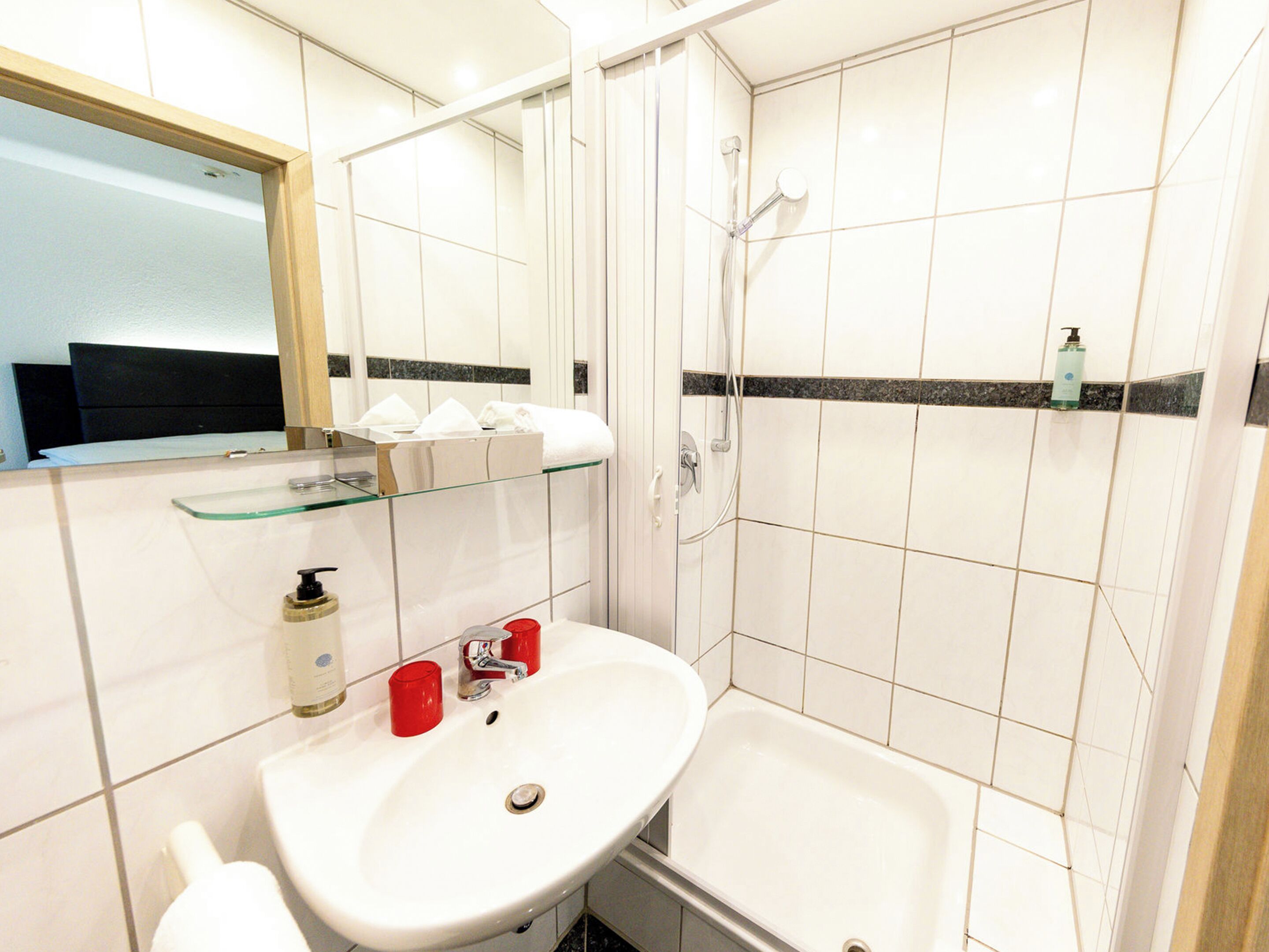 Un baño con lavabo, espejo, ducha y estante de vidrio con artículos de aseo.