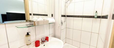Un baño con lavabo, espejo, ducha y estante de vidrio con artículos de aseo.