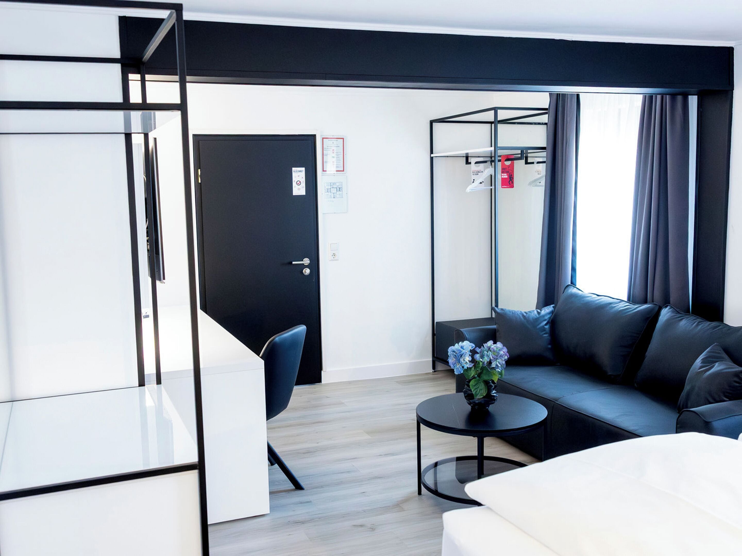 A modern hotel room with a black-framed bed, a white sofa, a small round table with a vase, and a black chair.