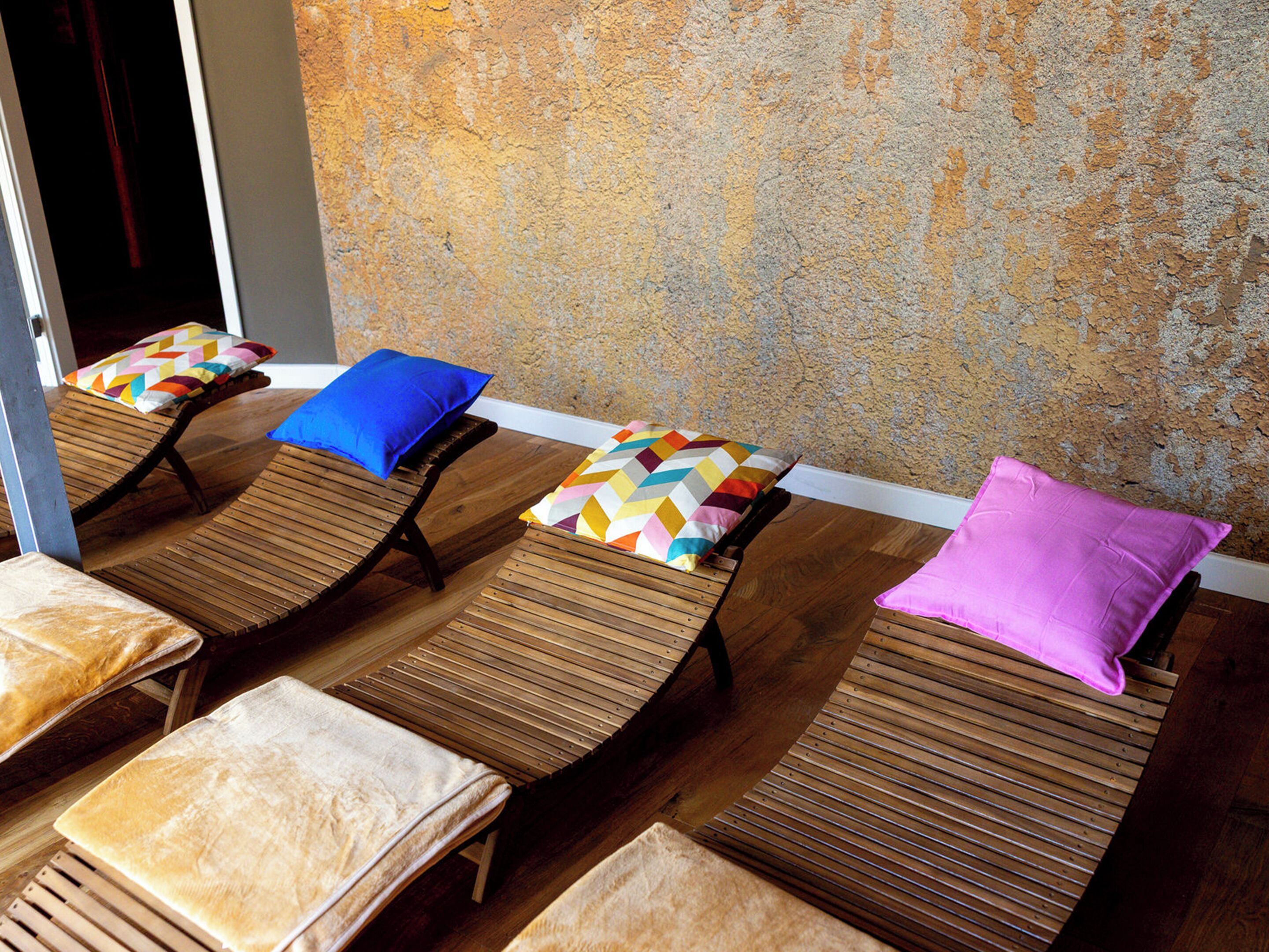 A row of wooden lounge chairs with colorful pillows against a textured wall.