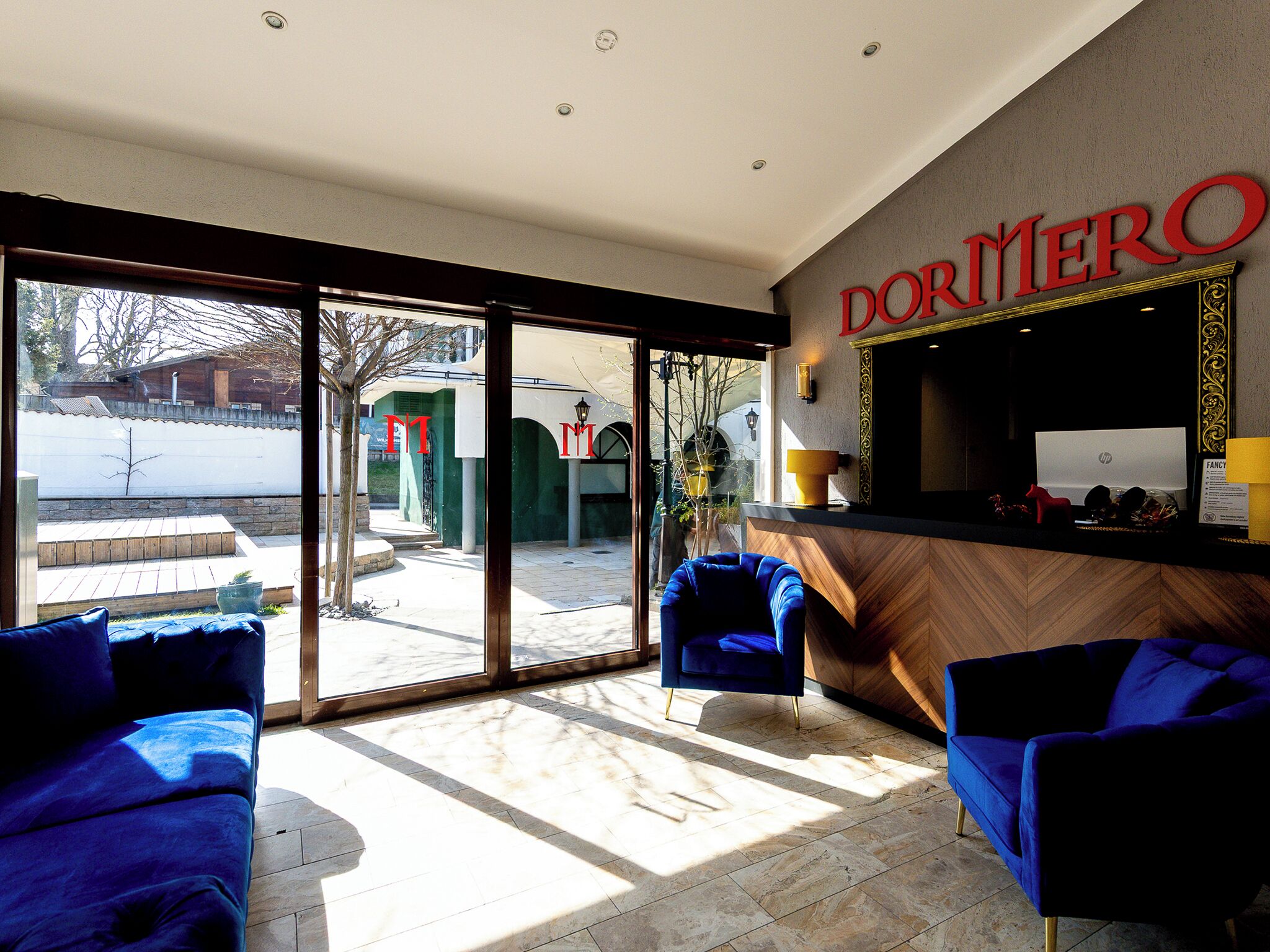 A hotel lobby with a reception desk, two blue armchairs, and a large glass door leading outside.