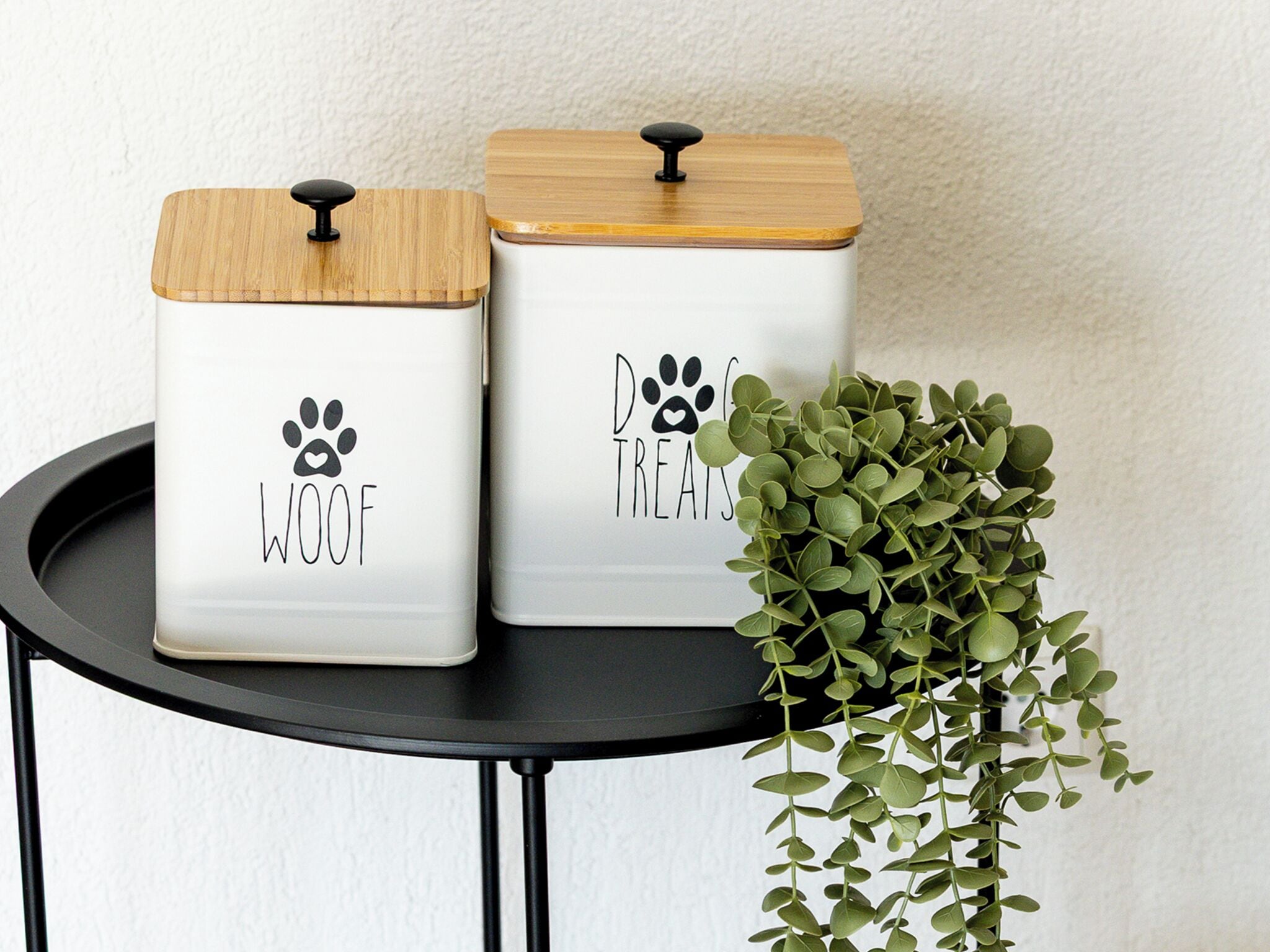 Two white storage containers with wooden lids and black handles, labeled 'WOOF' and 'DREAMS' respectively, on a black round table next to a green plant.