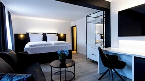 A modern hotel room with a bed, a desk, a chair, a small table with a vase, and a large mirror. - DORMERO Hotel Aalen (Aalen)