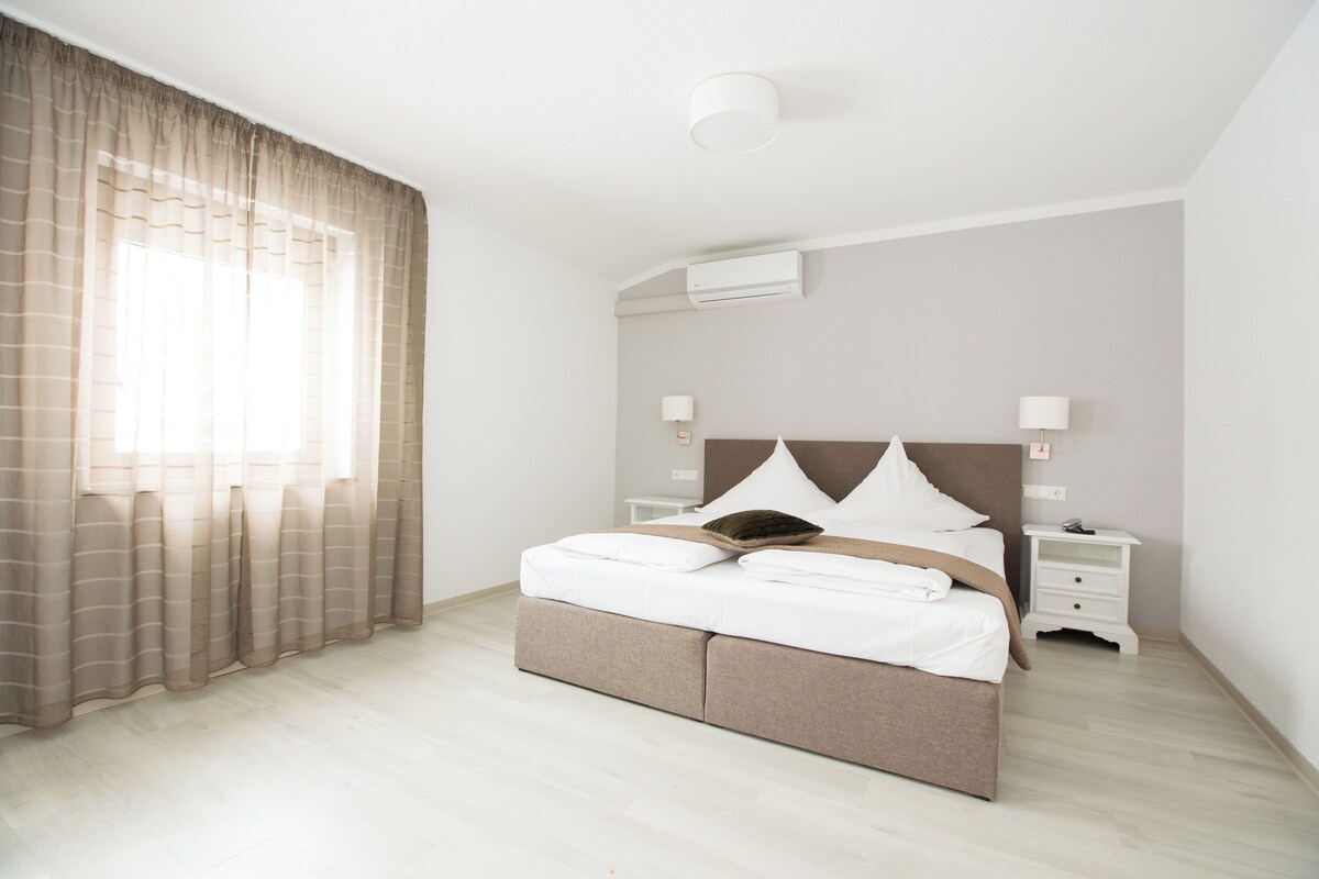 A neatly made bed with white linens and a brown headboard, two bedside tables with lamps, and a window with sheer curtains.