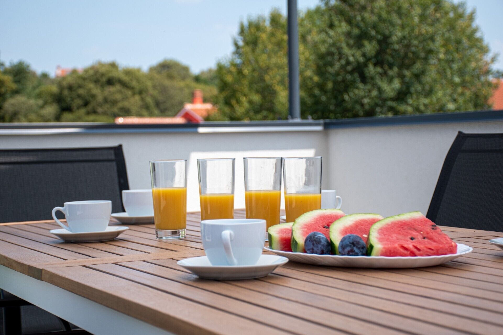 Une table en bois sur laquelle se trouvent une tasse blanche, deux verres de jus d’orange et une assiette de pastèque et de myrtilles.