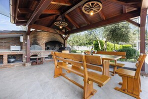An outdoor patio with a brick oven, wooden benches, and a table with a bottle and fruit.