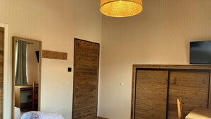 A bedroom with a bed, a wooden wardrobe, a desk, a mirror, and a television on the wall.