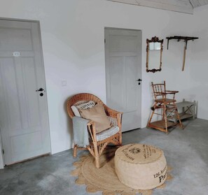 Quarto com duas portas, uma cadeira de vime com almofada, uma cadeira de madeira e uma mesa redonda com etiqueta.