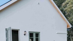 A white building with a wooden deck, a dining area with chairs and a table, a window with green shutters, and a potted plant with flowers. - Cozy house in small village near the sea (Skåne län)