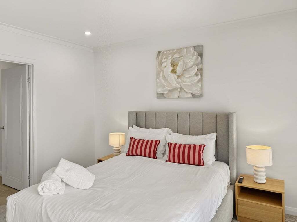 A neatly made bed with white bedding and red striped pillows, a grey upholstered headboard, and a wall-mounted abstract painting.