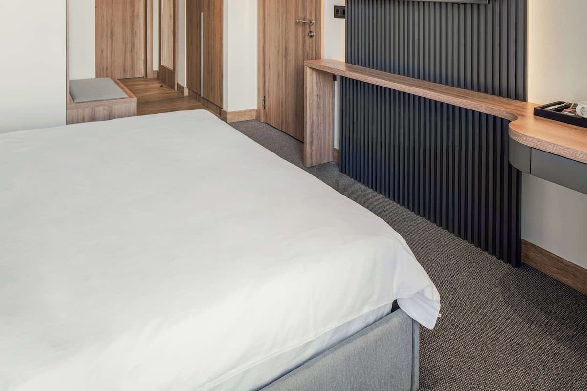 A hotel room with a large bed, a flat-screen TV mounted on a dark wall, a wooden bench, and a small seating area.