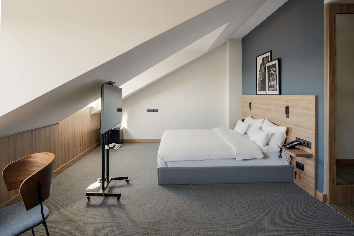 A modern hotel room with a sloped ceiling, a large bed, a desk, and a chair.