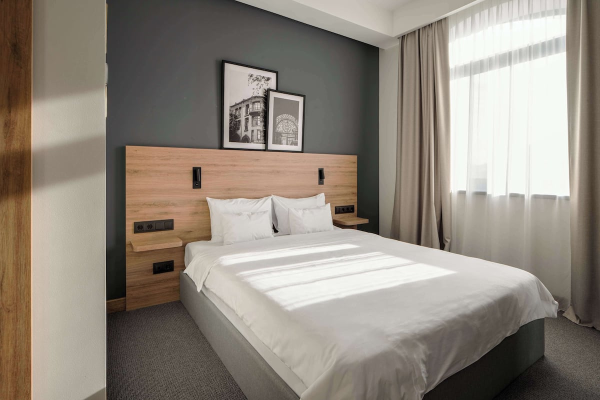 A hotel room with a large bed, wooden headboard, two framed pictures, a nightstand, and a window with curtains.