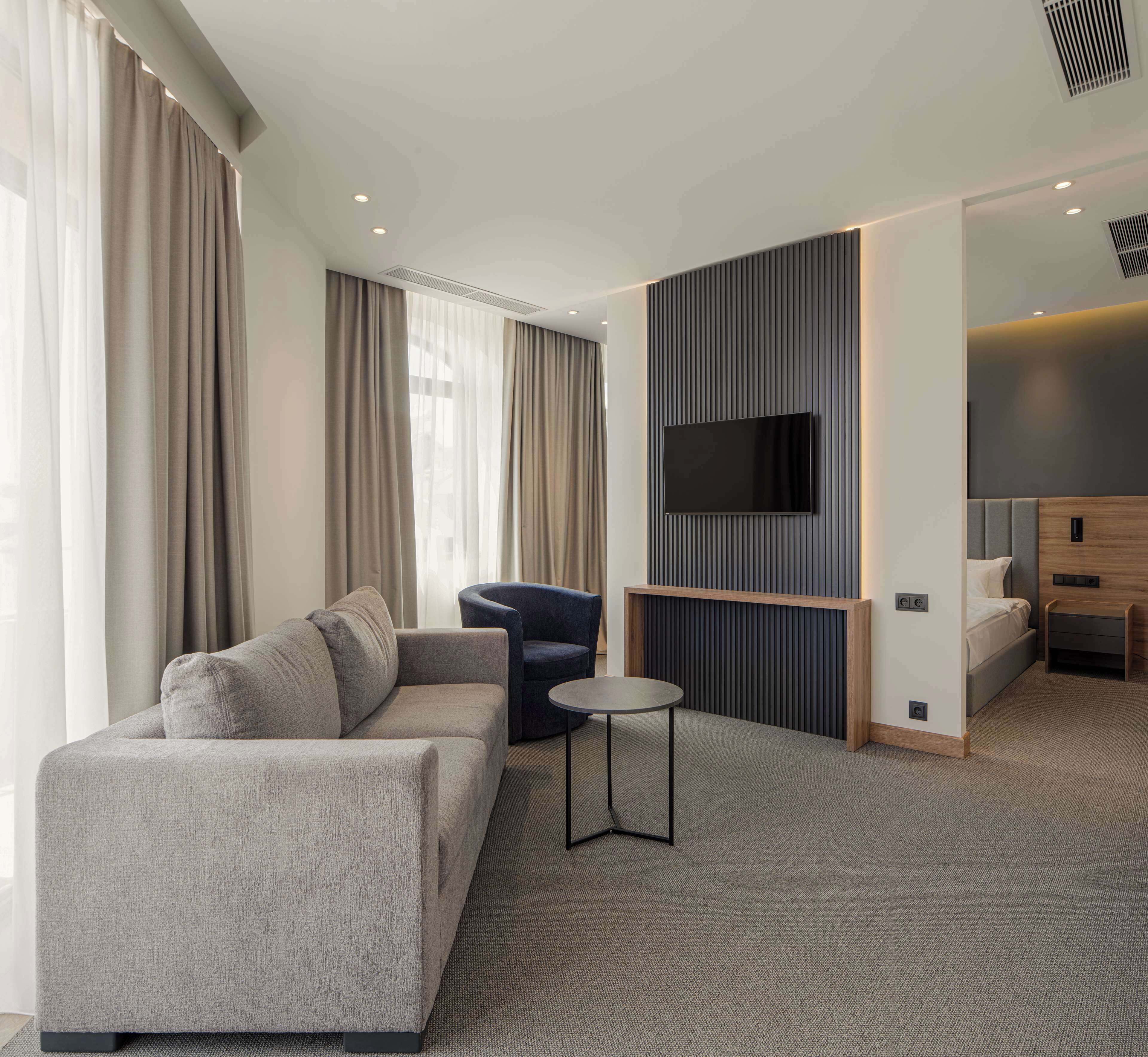 A modern hotel room with a grey sofa, a small round table, a blue armchair, a TV mounted on a dark panel wall, and a bed with a wooden headboard.