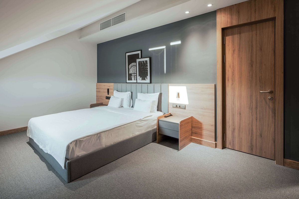 A modern hotel room with a large bed, a bedside table with a lamp, and a wooden wardrobe.