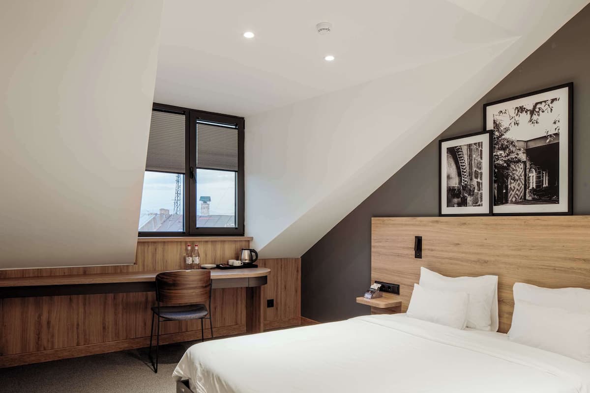 A modern bedroom with a sloped ceiling, a large bed, a desk with a chair, and two framed pictures on the wall.