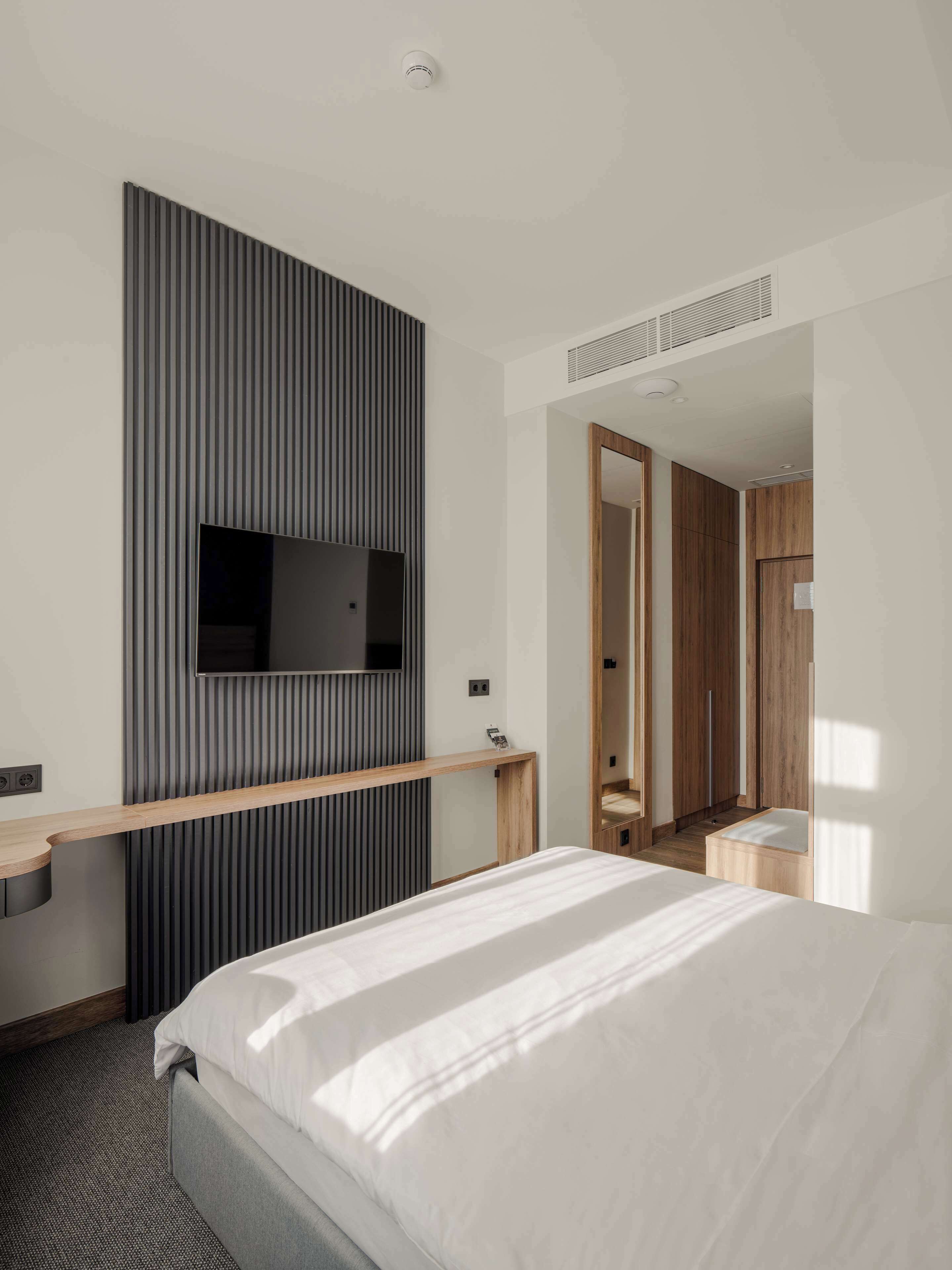 A modern hotel room with a bed, a mounted TV, a wooden console table, and a mirror.