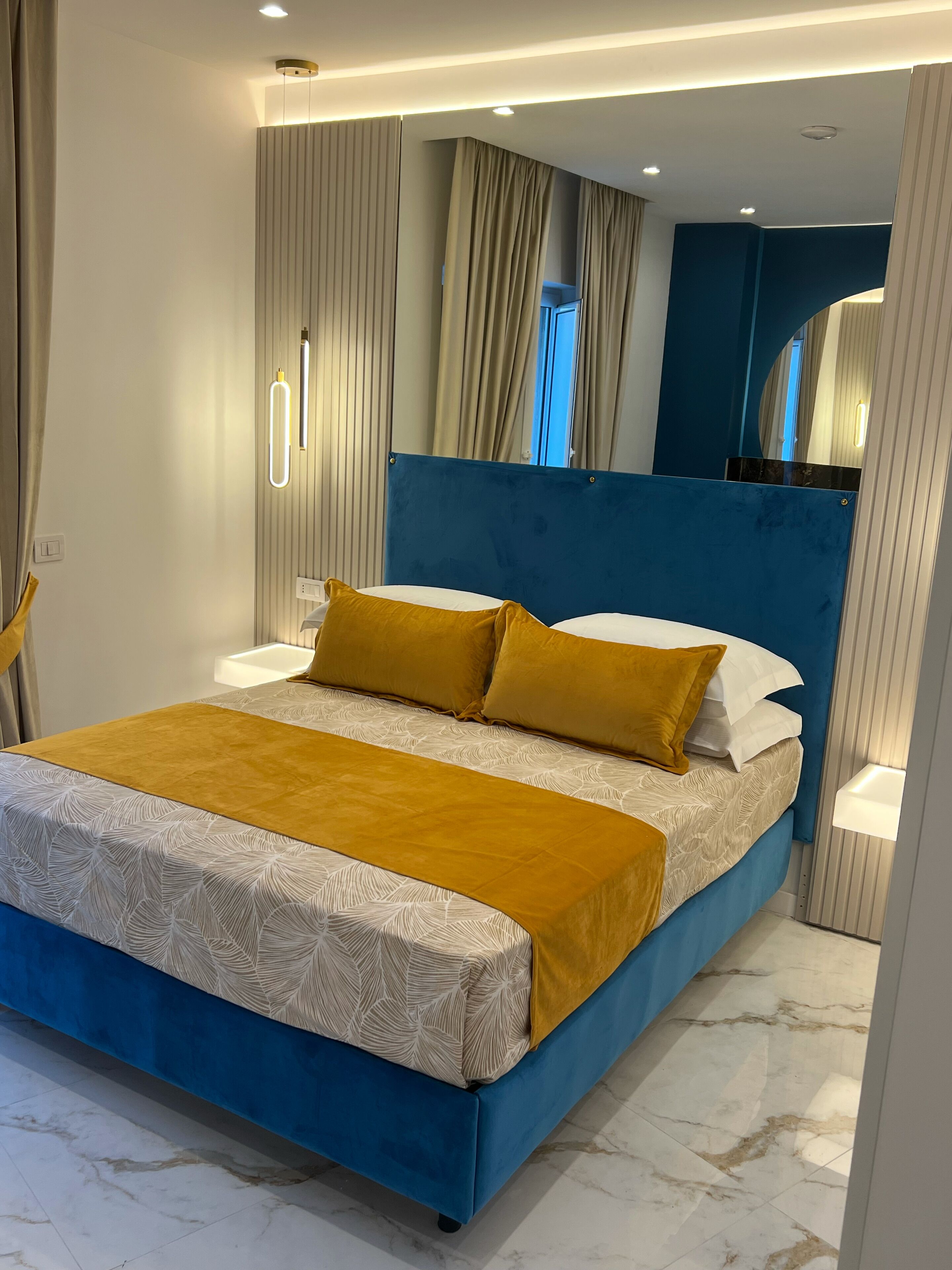 Een moderne slaapkamer met een blauw hoofdeinde, een groot bed met wit en geel beddengoed, en een marmeren vloer.