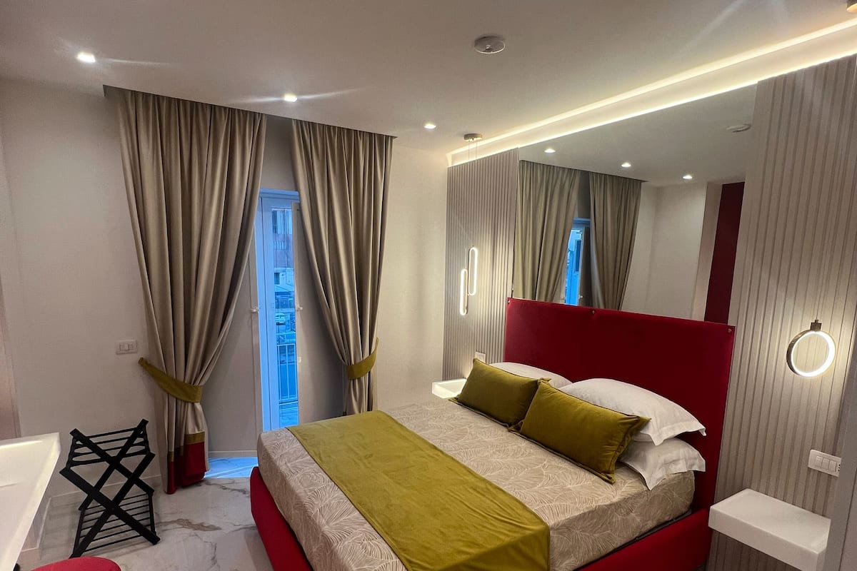 Un dormitorio moderno con cama roja, una mesita de noche pequeña, una silla roja y un baño con espejo y lavamanos.