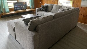 Un salon chaleureux avec un canapé d’angle gris, un téléviseur à écran plat posé sur un meuble en bois et une fenêtre donnant sur l’extérieur.