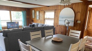 L’intérieur chaleureux d’un chalet, avec un décor en bois, un canapé gris, une table à manger avec des chaises et une grande fenêtre donnant sur l’océan.