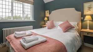 Un lit bien fait, avec du linge de lit blanc et des coussins roses, une fenêtre donnant sur un paysage verdoyant et un radiateur placé sous la fenêtre.