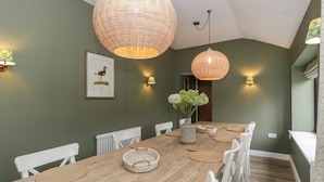 Un espace repas avec une longue table en bois, des chaises blanches, des sets de table en fibres tressées et deux grands luminaires suspendus en fibres tressées.