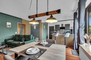 Ein modernes Wohnzimmer mit Essbereich, grünen Wänden, Holzelementen und einer Küche im Hintergrund.