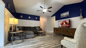 A living room with a dark blue accent wall, a ceiling fan, a sofa, a coffee table, a patterned armchair, and a television. - Foley Vacation or Monthly Stay Near OWA | Sleeps 12 (Foley)