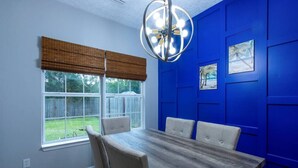 A dining room with a wooden table and chairs, a window with a view of a backyard, and a blue wall with paneling and framed pictures. - Foley Vacation or Monthly Stay Near OWA | Sleeps 12 (Foley)