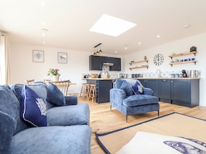 A modern living room with a sofa and armchair, a dining area with a table and chairs, and a kitchen with dark cabinets and wooden shelves. - Chi an Mor (Penzance)