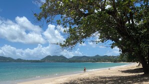 A beach with clear turquoise water, a sandy shore, and a person walking with a yellow surfboard. - Fabulous poolside apartment Marigot Bay. (Marigot bay)