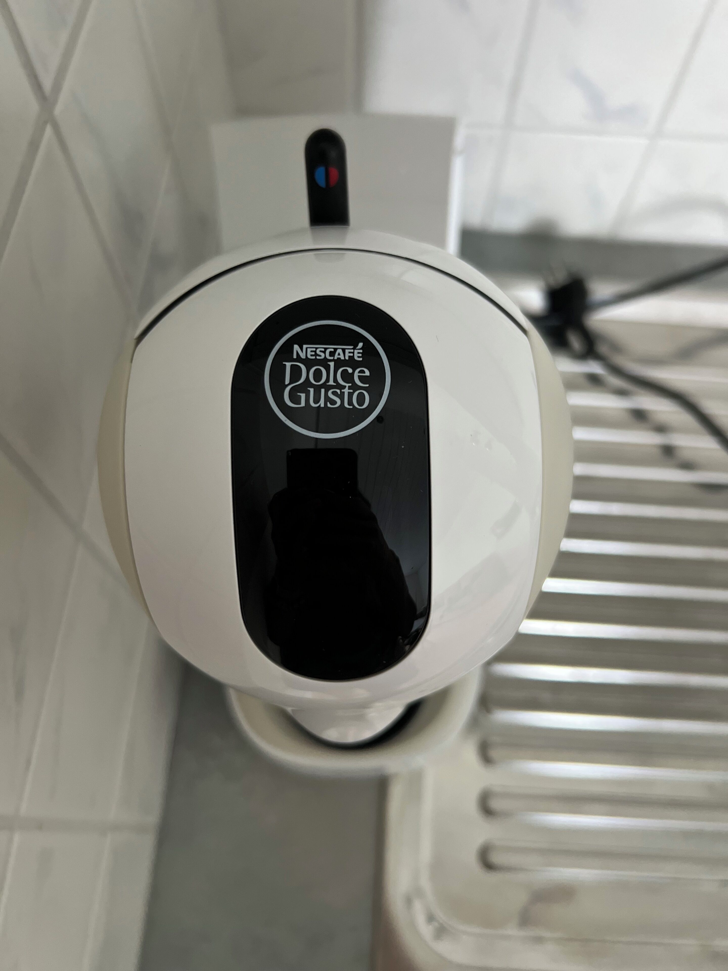 En vit Nescafé Dolce Gusto-kaffemaskin monterad på en vägg, med en kakelbeklädd vägg bakom.