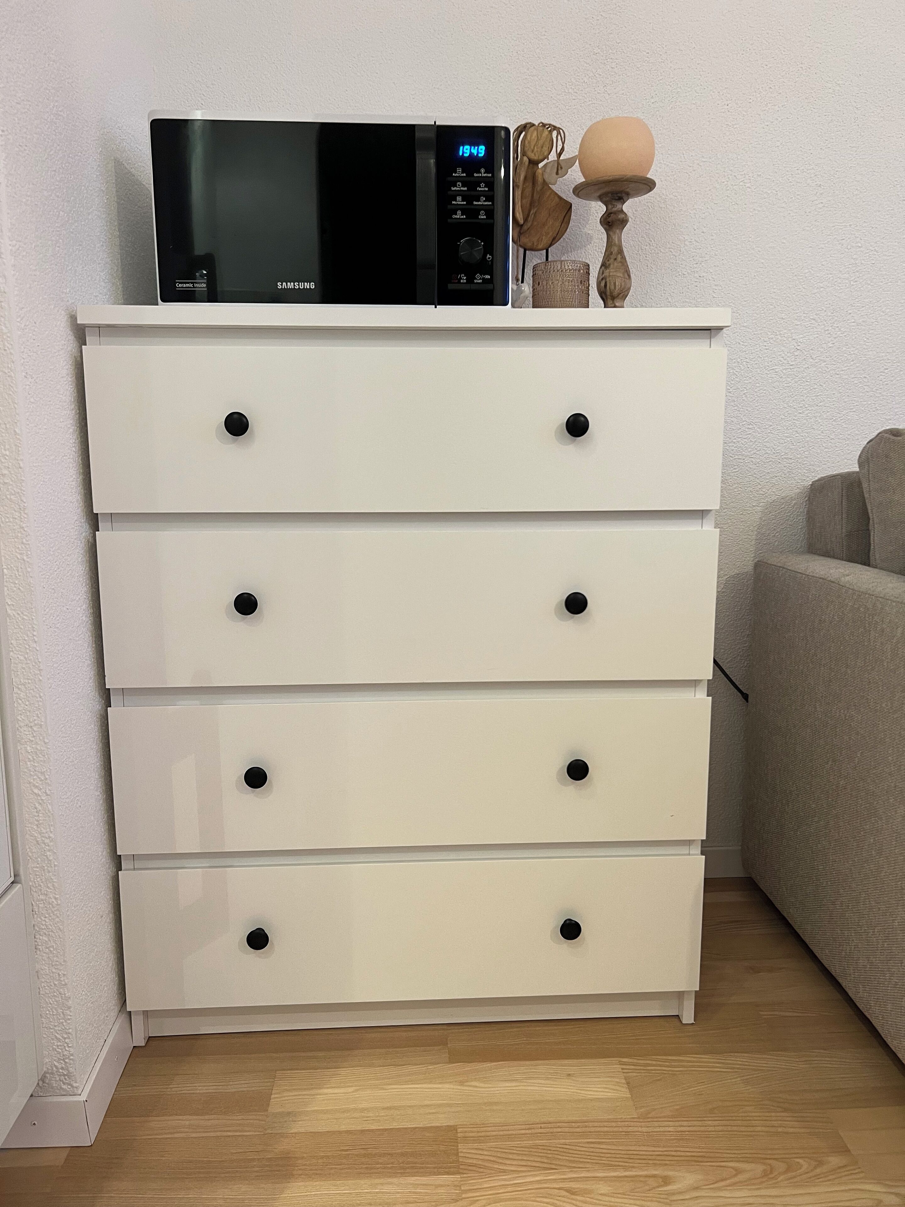 A white chest of drawers with black knobs, a black microwave on top, and a small decorative item beside it.
