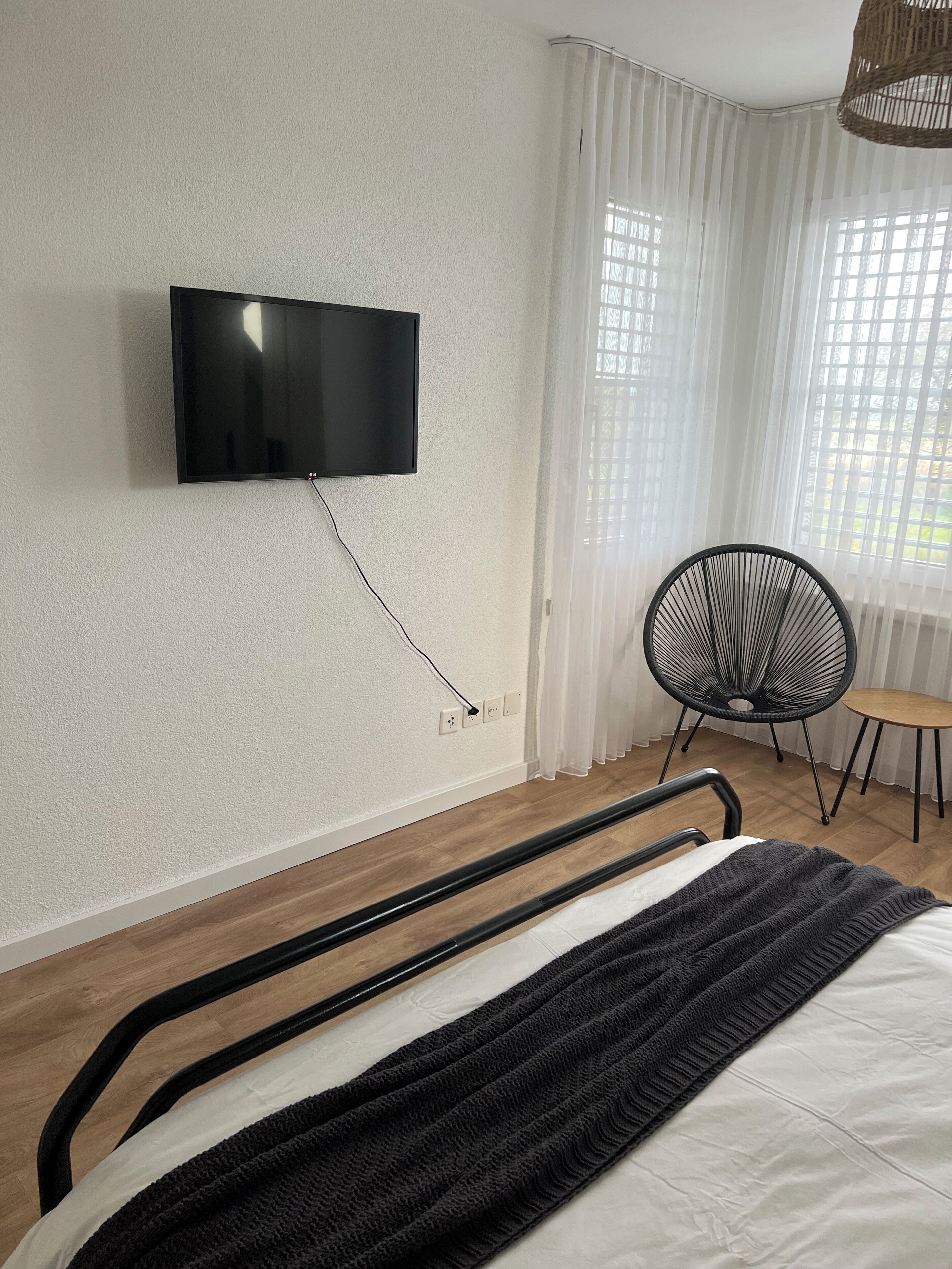 Une chambre à coucher avec un lit, un téléviseur fixé au mur, une chaise et une fenêtre avec des rideaux.