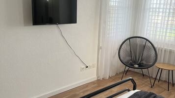 Une chambre à coucher avec un lit, un téléviseur fixé au mur, une chaise et une fenêtre avec des rideaux.