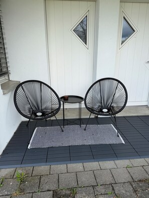 Eine Terrasse mit zwei schwarzen Drahtstühlen, einem kleinen Tisch, einem gemusterten Teppich und einer weißen Tür mit Fenstern.