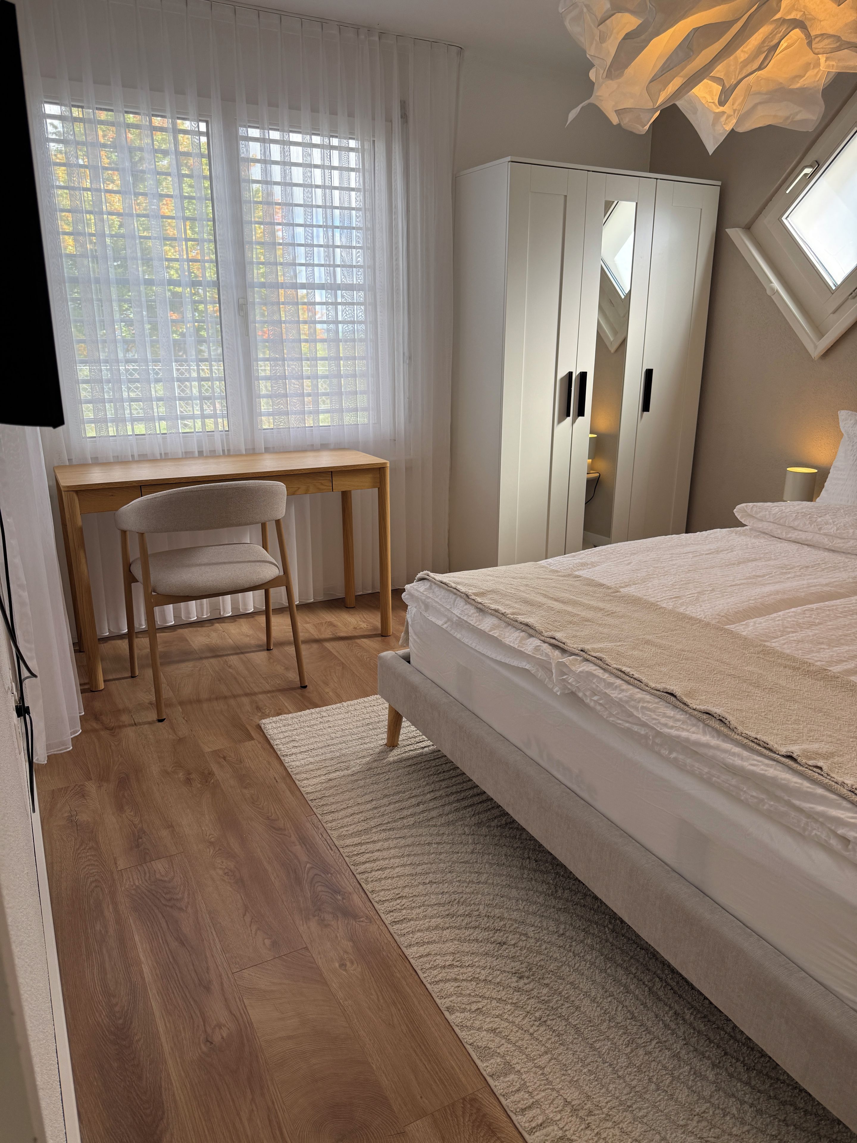 A bedroom with a bed, a desk, a chair, a wardrobe, and a window with curtains.
