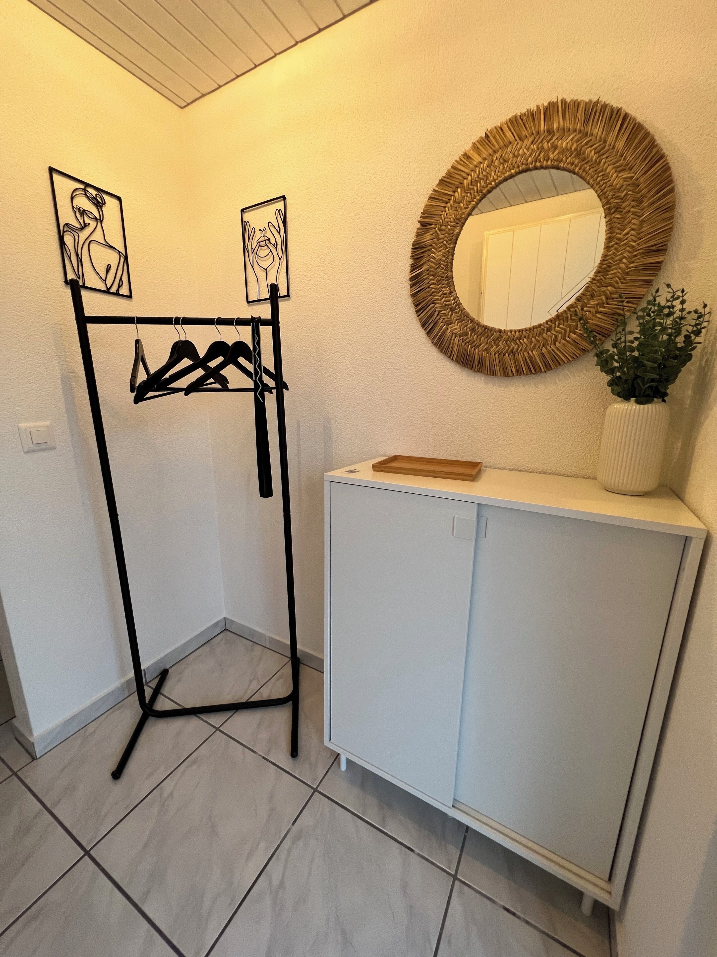 Une pièce avec un meuble blanc, un étendoir à linge noir en métal et un miroir rond doté d’un cadre tressé.
