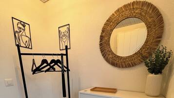Une pièce avec un meuble blanc, un étendoir à linge noir en métal et un miroir rond doté d’un cadre tressé.