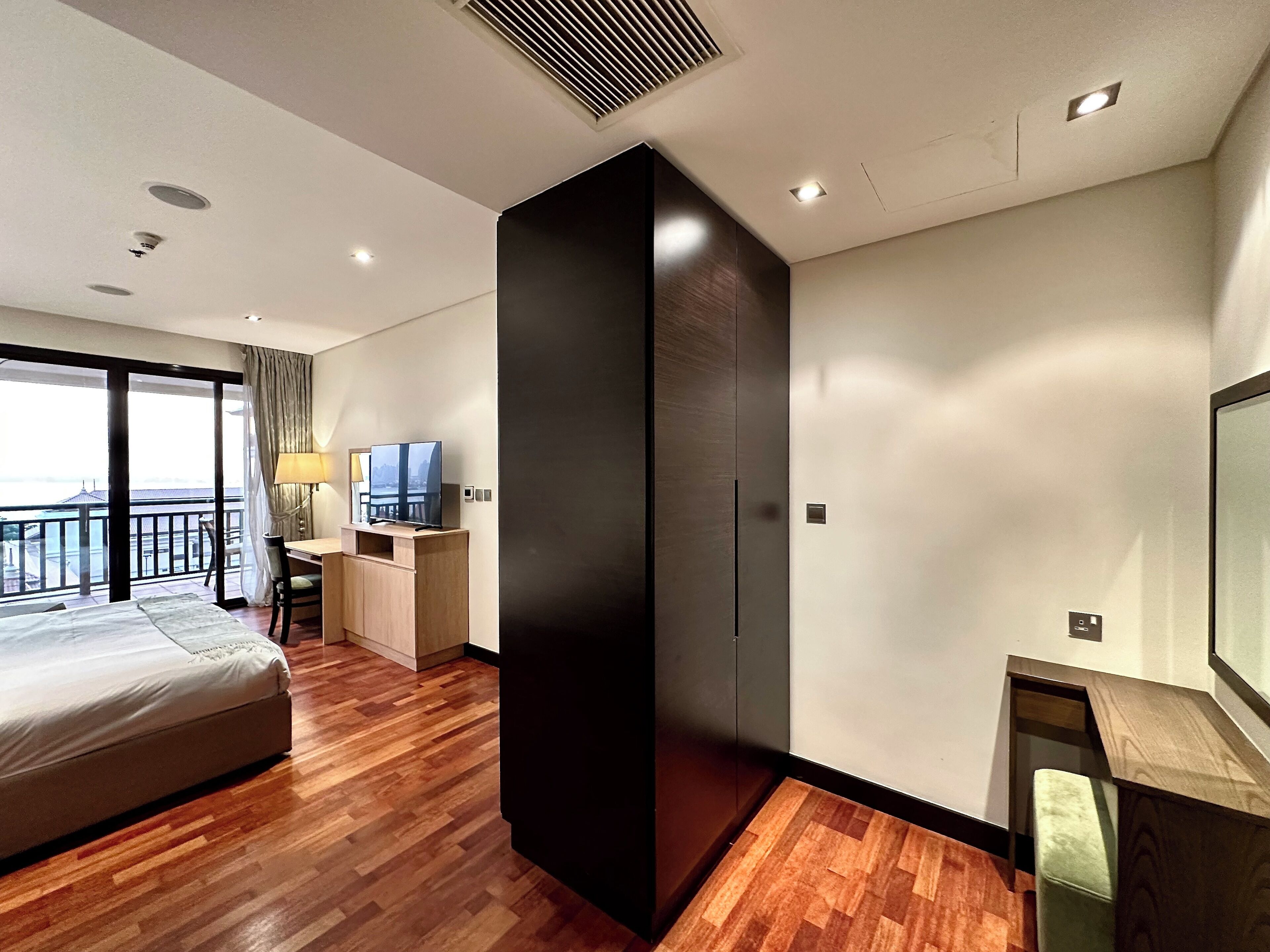 A modern hotel room with a large bed, a desk with a TV, a wardrobe, and a balcony with a view.