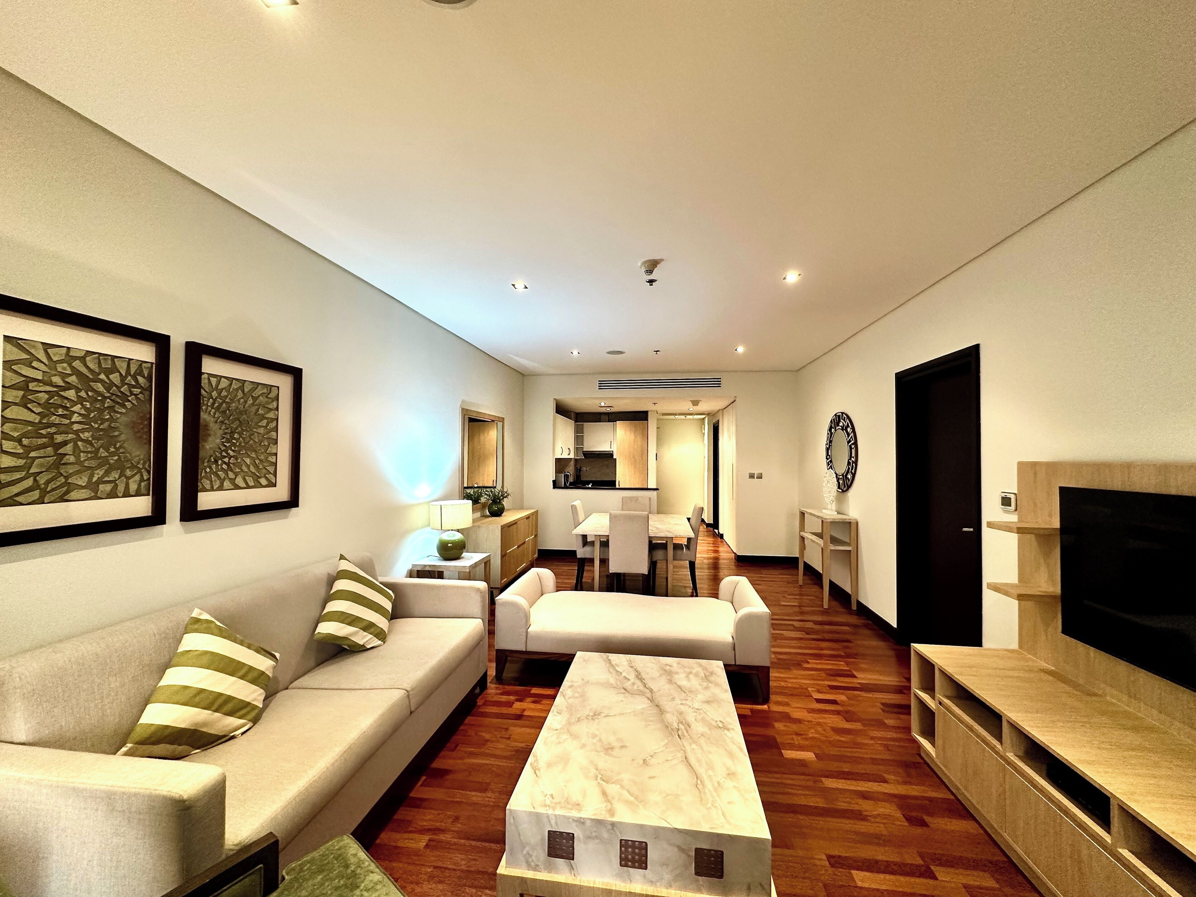 A modern living room with a sofa, a coffee table, and a TV stand. There is a dining area with a table and chairs in the background.