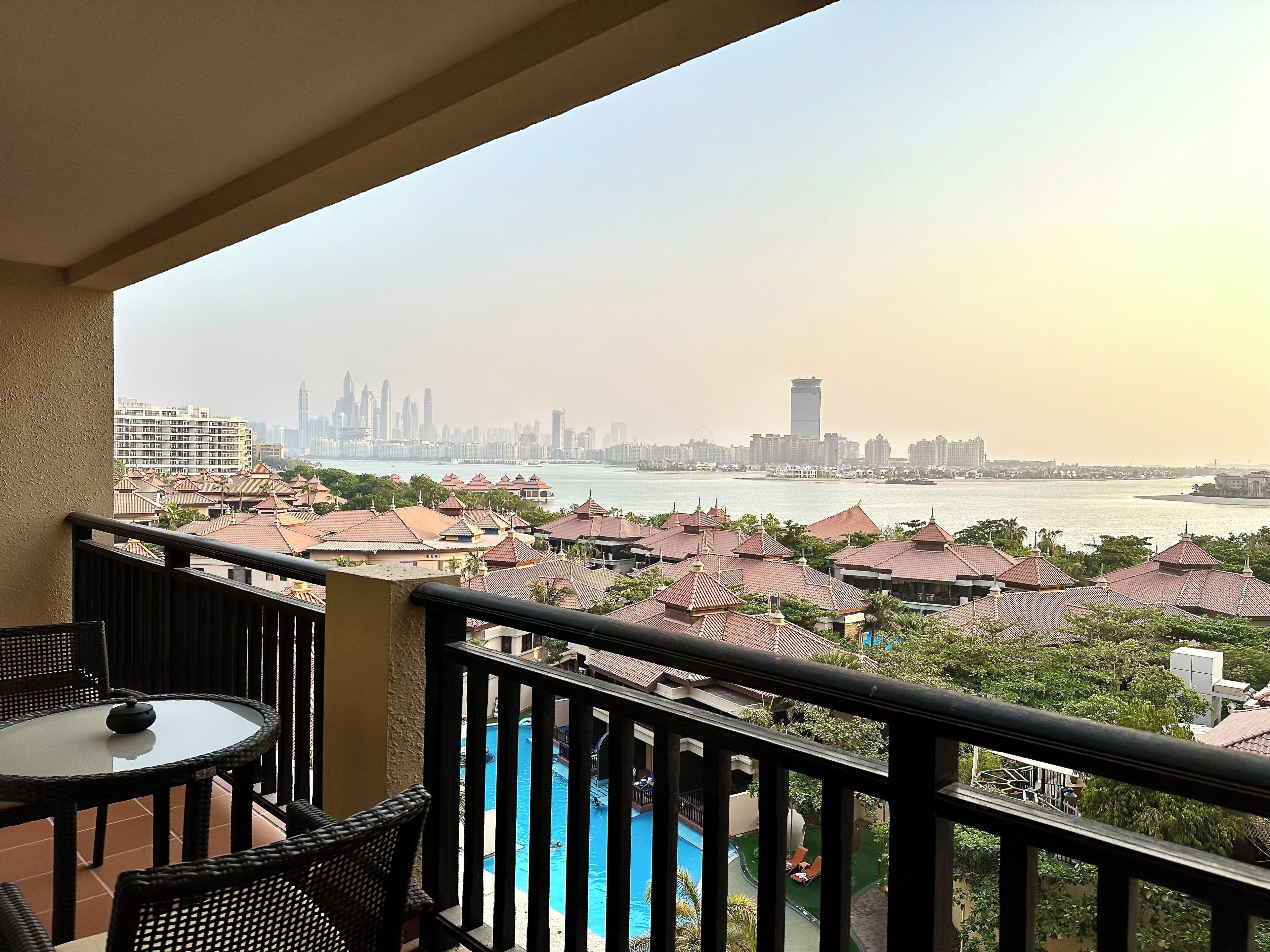 A balcony with a view of a city skyline, a swimming pool, and a distant body of water.