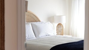 Un lit bien fait, avec des draps blancs et une couverture bleu foncé, une tête de lit en osier et une lampe de chevet avec un abat-jour blanc.