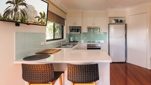 Une cuisine moderne avec des meubles blancs, un îlot central doté de deux tabourets de bar, un four à micro-ondes, un réfrigérateur et une grande fenêtre offrant une vue sur les palmiers.